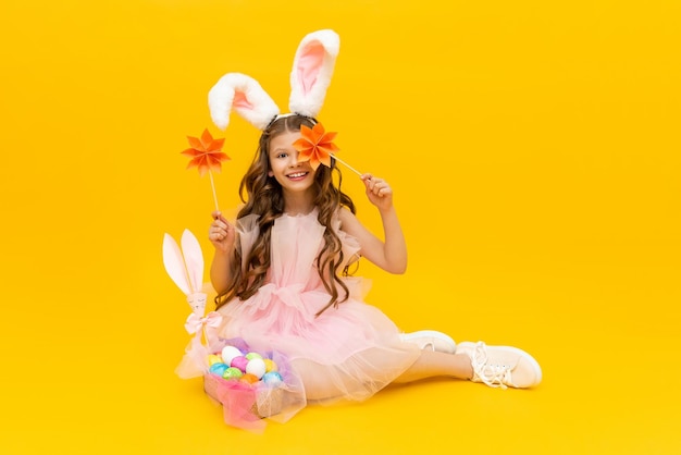 Festive Easter A curlyhaired girl with rabbit ears in a pink dress is enjoying the holiday A child with a basket of colorful eggs on a yellow isolated background