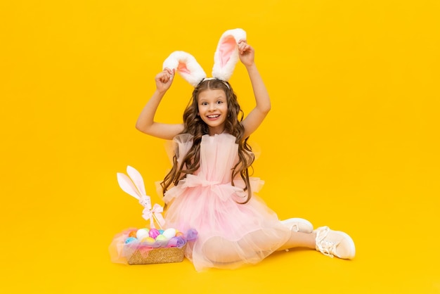 Festive Easter A curlyhaired girl with rabbit ears in a pink dress is enjoying the holiday A child with a basket of colorful eggs on a yellow isolated background