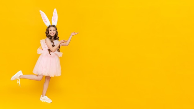 Festive Easter A teenage girl in a fluffy dress and with rabbit ears points to your advertisement on a yellow isolated background A child in a rabbit costume stands tall Copy space