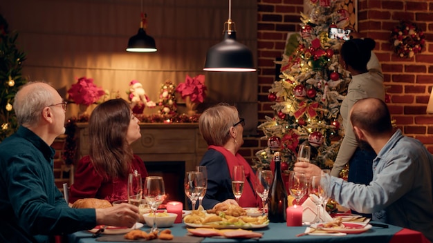 Festive family taking selfie photos with smartphone device while sitting at Christmas dinner table. Happy diverse people celebrating winter feast while taking photos of themselves.