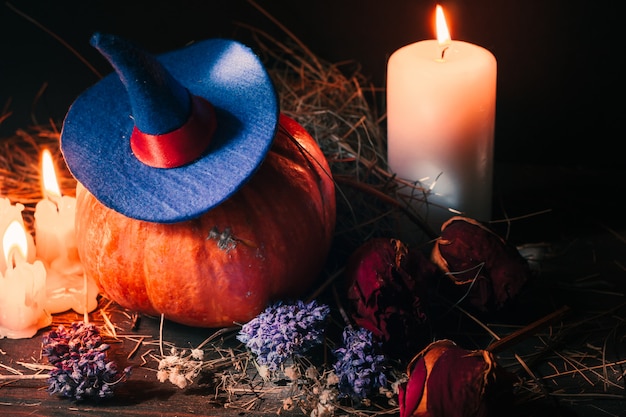 Festive Halloween decoration with orange pumpkin with witch hat, candles, roses and dry hay.