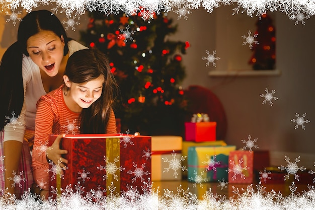 Festive mother and daughter opening a glowing christmas gift against fir tree forest and snowflakes