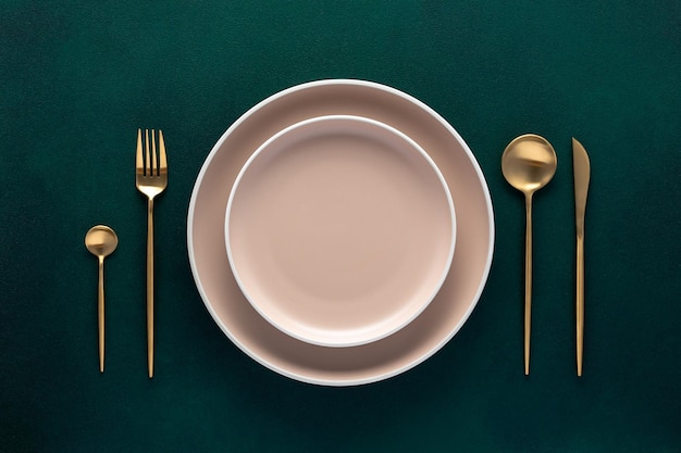 Festive place setting with beige dishes Empty plate and gold cutlery on dark green background Dining table in luxury restaurant Card or menu template flat design Tableware crockery Top view