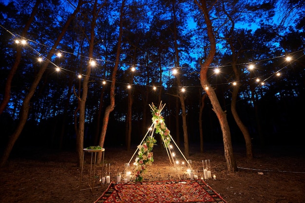 Festive string lights illumination on boho tipi arch decor on outdoor wedding ceremony venue in pine