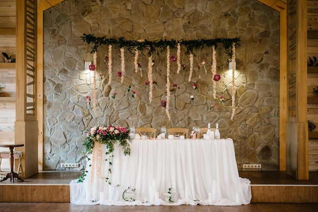 Festive table newlyweds covered with a tablecloth and decorated with composition of flowers and greenery candles in the wedding banquet hall Wedding party in tent with wooden and stone wall