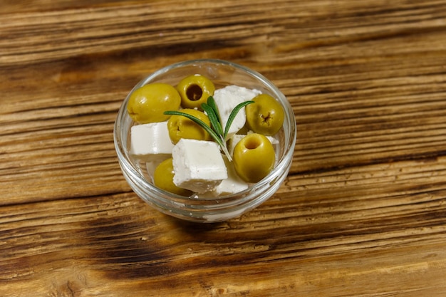 Feta cheese cubes with green olives and rosemary in glass bowl on a wooden table