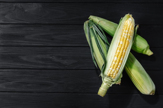 A few corn in leaves lie on a wooden dark background view from above space for text