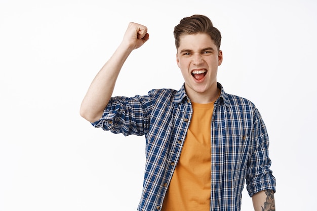 Fierce and excited young man chanting, fist pump and yelling, watching competition, sports game fan looking at tv, shouting with joy, celebrating, standing on white