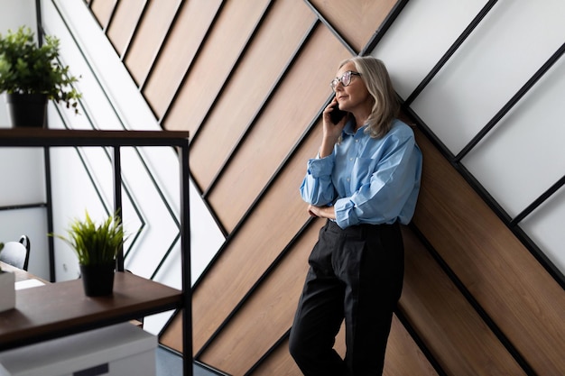 Fifty year old successful business woman executive talking on mobile phone leaning against the wall