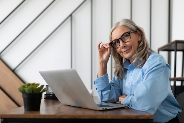 Fiftyyearold businesswoman sits in front of a laptop at a table in the office and adjusts her