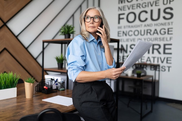 Fiftyyearold businesswoman with documents in her hands speaks on a mobile phone in a modern office