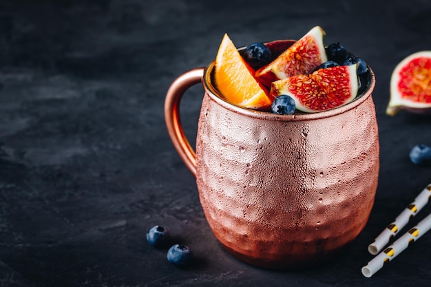 Fig moscow mule ice cold cocktail in copper cup with blueberries and orange on dark stone background