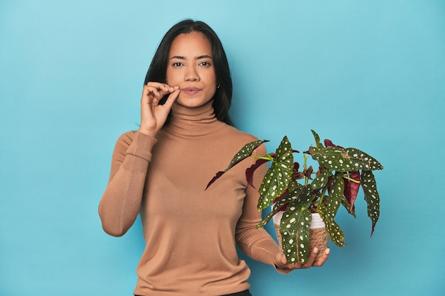 Filipina holding a plant on blue studio with fingers on lips keeping a secret