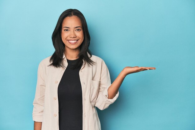 Photo filipina young woman on blue studio showing a copy space on a palm and holding another hand on waist