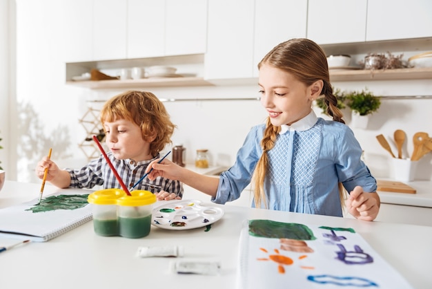 Final touch. Attentive creative beautiful lady adding some details to the masterpiece of her sibling while he looking away and they both spending the morning enjoying their common hobby