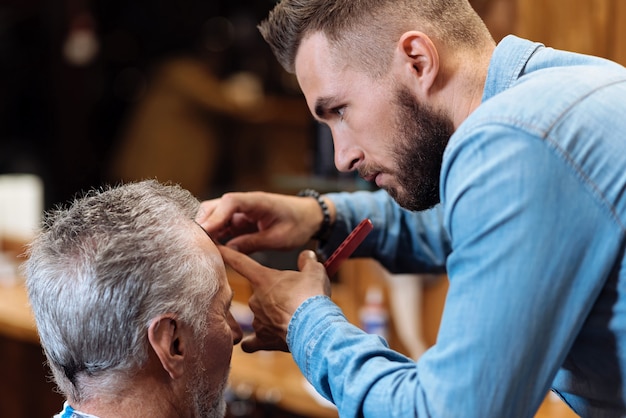 Final touch. Close up of young handsome barber concentrated doing haircut to senior man at barbershop.