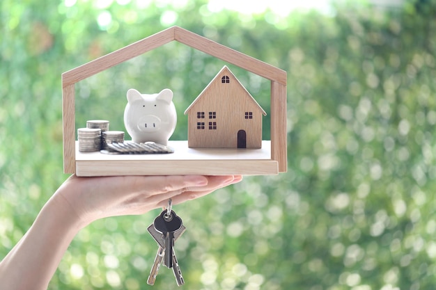 Finance Woman hand holding model house with piggy bank and stack of coins money on natural green backgroundBusiness investment and real estate concept