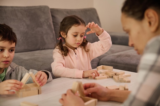 Fine motor skills, concentration development, educational leisure, family pastime concept. Cute kids playing board game with their blurred mom focused on building a wooden structure in the foreground