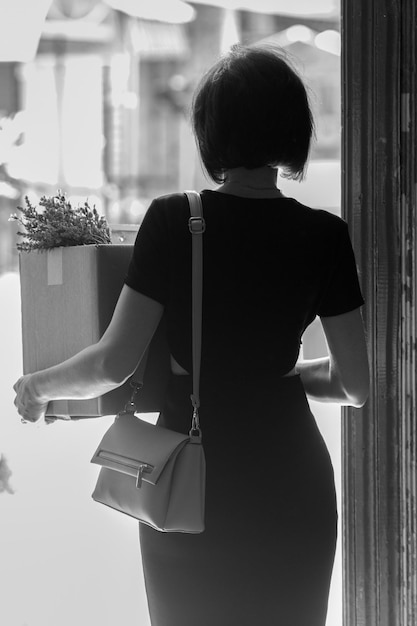 A fired brunette in a black dress with a box of personal belongings. Black and white