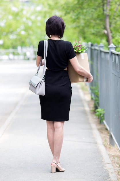 A fired brunette in a black dress with a box of personal belongings.