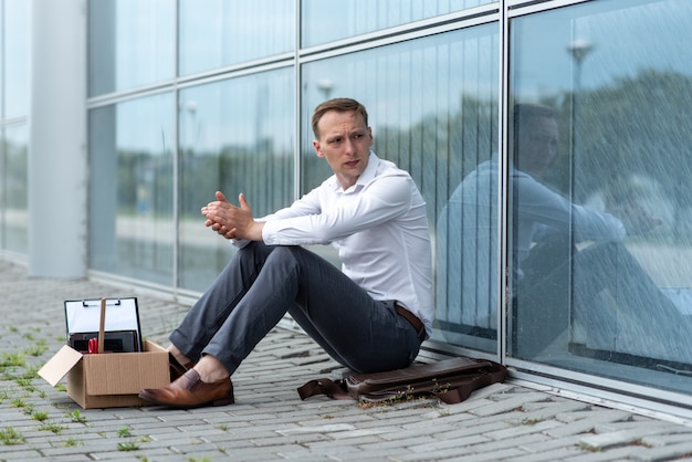 A fired office worker sits on the floor near a modern office building. The man is very worried about the dismissal. The man looks away and shakes hands.