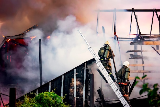 Firefighters battle a wildfire against a background of thick white smoke.. Firefighters on the stairs extinguish the roof of a private house or barn.