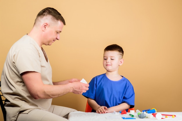 First aid. Medicine concept. Kid little doctor sit table medical tools. Health care. Medical examination. Boy cute child and his father doctor. Hospital worker. Medical help. Medical insurance.