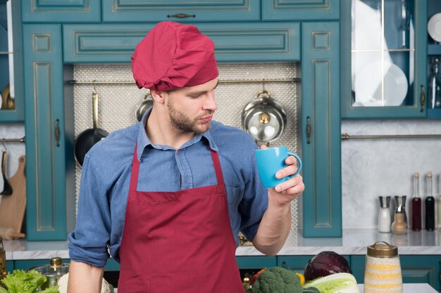 But first coffee Man with holds coffee cup Cook stands near table with ingredients and kitchenware Self care Cooking process concept Chef in burgundy uniform drink coffee for inspiration