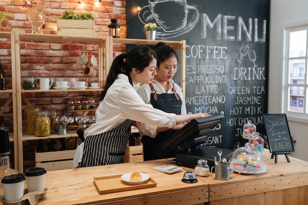 first part time job in summer concept. professional barista teaching young girl new employee how to using tablet to take customer order in cafe bar counter in coffee shop. coworkers help each other