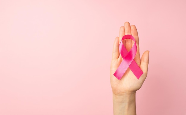 First person top view photo of female hand holding pink ribbon in palm symbol of breast cancer awareness on isolated pastel pink background with copyspace