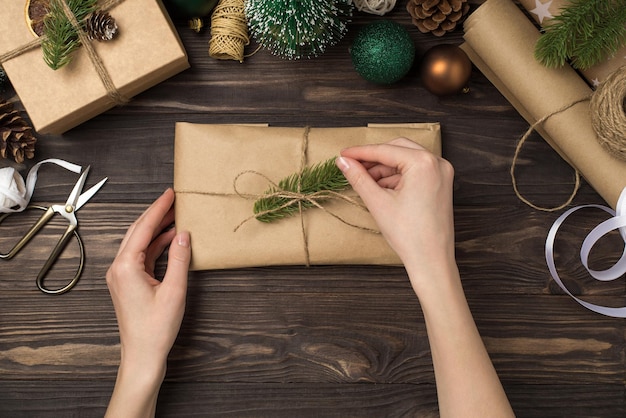 First person top view photo of female hands decorating craft paper giftbox with pine twigs and twine handicraft tools cones green and gold christmas tree balls on isolated wooden desk background