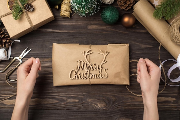 First person top view photo of female hands decorating kraft paper package with merry christmas wooden text and twine green gold balls pine twigs handicraft tools on isolated wooden desk background