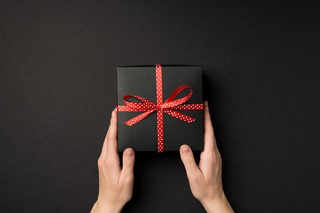 First person top view photo of female hands holding black giftbox with red dotted ribbon bow on isolated black background