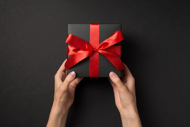 First person top view photo of female hands holding black giftbox with vivid red ribbon bow on isolated black background