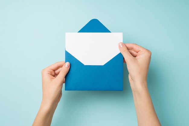 First person top view photo of female hands holding open blue envelope with white paper sheet on isolated pastel blue background with empty space