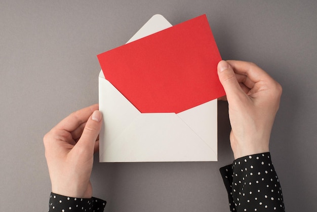 First person top view photo of female hands holding open white envelope with red card on isolated grey background with copyspace