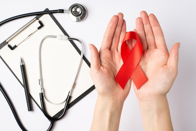 First person top view photo of female hands holding red ribbon in palms over clipboard pen and stethoscope symbol of aids awareness on isolated white background