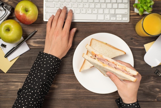 First person top view photo of female hands holding sandwich over plate and typing on keyboard mouse apples glass of juice flowerpot and stationery on isolated dark wooden table background