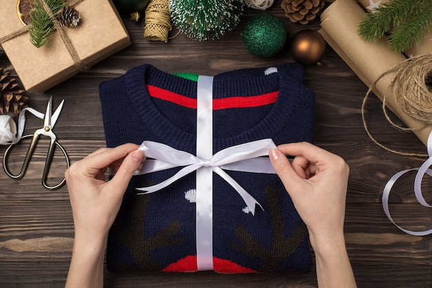 First person top view photo of female hands tying ribbon bow on folded sweater craft paper giftbox green gold christmas tree balls pine twigs and handicraft tools on isolated wooden desk background