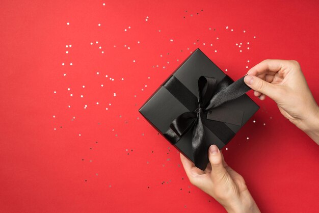 First person top view photo of female hands unpacking stylish black giftbox with black ribbon bow and tag on isolated red background with blank space