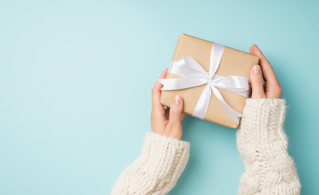 First person top view photo of female hands in white cozy sweater holding craft paper giftbox with white satin ribbon bow on isolated pastel blue background with copyspace