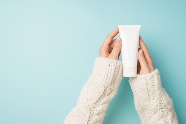 First person top view photo of female hands in white sweater holding white cream tube without label on isolated pastel blue background with blank space