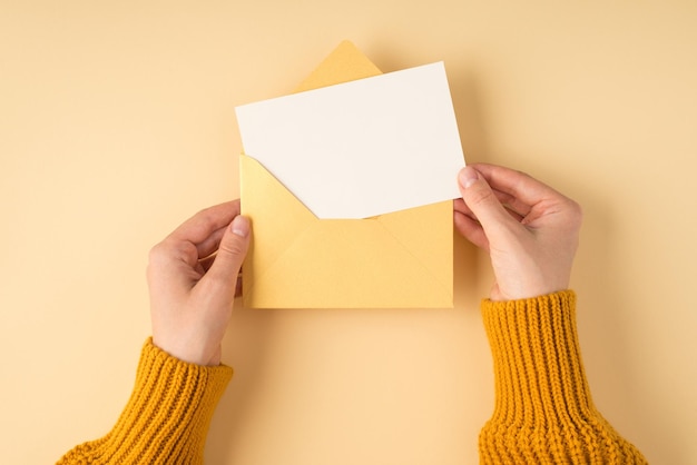 First person top view photo of female hands in yellow pullover holding open pastel yellow envelope with white card on isolated light orange background with copyspace