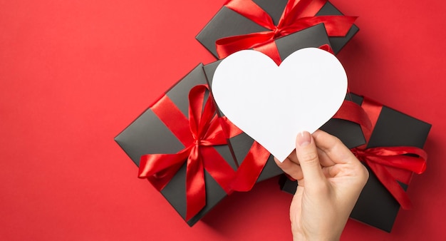 First person top view photo of hand holding white paper heart over stack of gift boxes on isolated red background with blank space