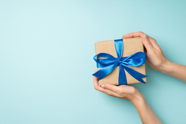 First person top view photo of hands demonstrating craft paper giftbox with vivid blue satin ribbon bow on isolated pastel blue background with copyspace