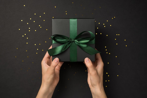 First person top view photo of hands giving black gift box with green ribbon bow and golden confetti on isolated black background