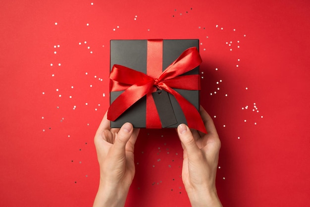 First person top view photo of hands holding black giftbox with red ribbon bow and tag shiny sequins on isolated red background with blank space on pricetag