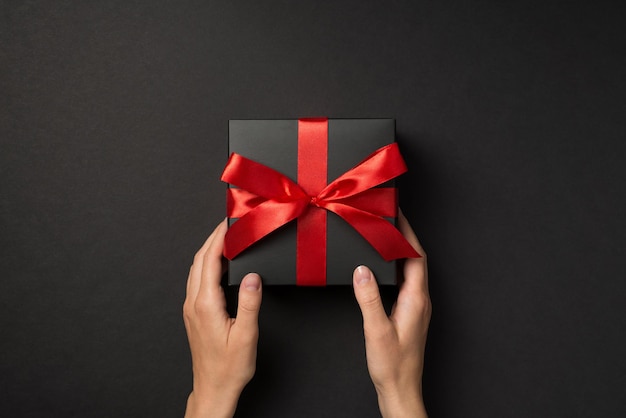 First person top view photo of hands holding black giftbox with vivid red ribbon bow on isolated black background