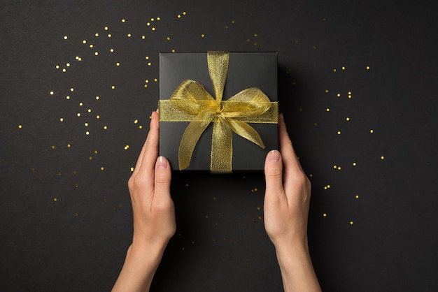 First person top view photo of hands holding black giftbox with yellow mesh ribbon bow over golden sequins on isolated black background