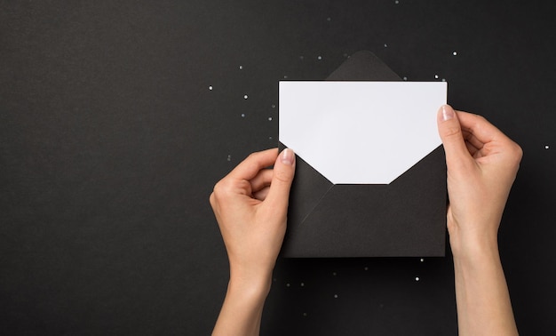 First person top view photo of hands holding black open envelope with white card over sequins on isolated black background with blank space